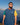 Man wearing a colorful t-shirt with a logo, standing by a body of water with a cityscape in the background.