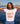 Woman wearing a white t-shirt with red accents and a graphic design, standing by a waterfront with a city skyline in the background.